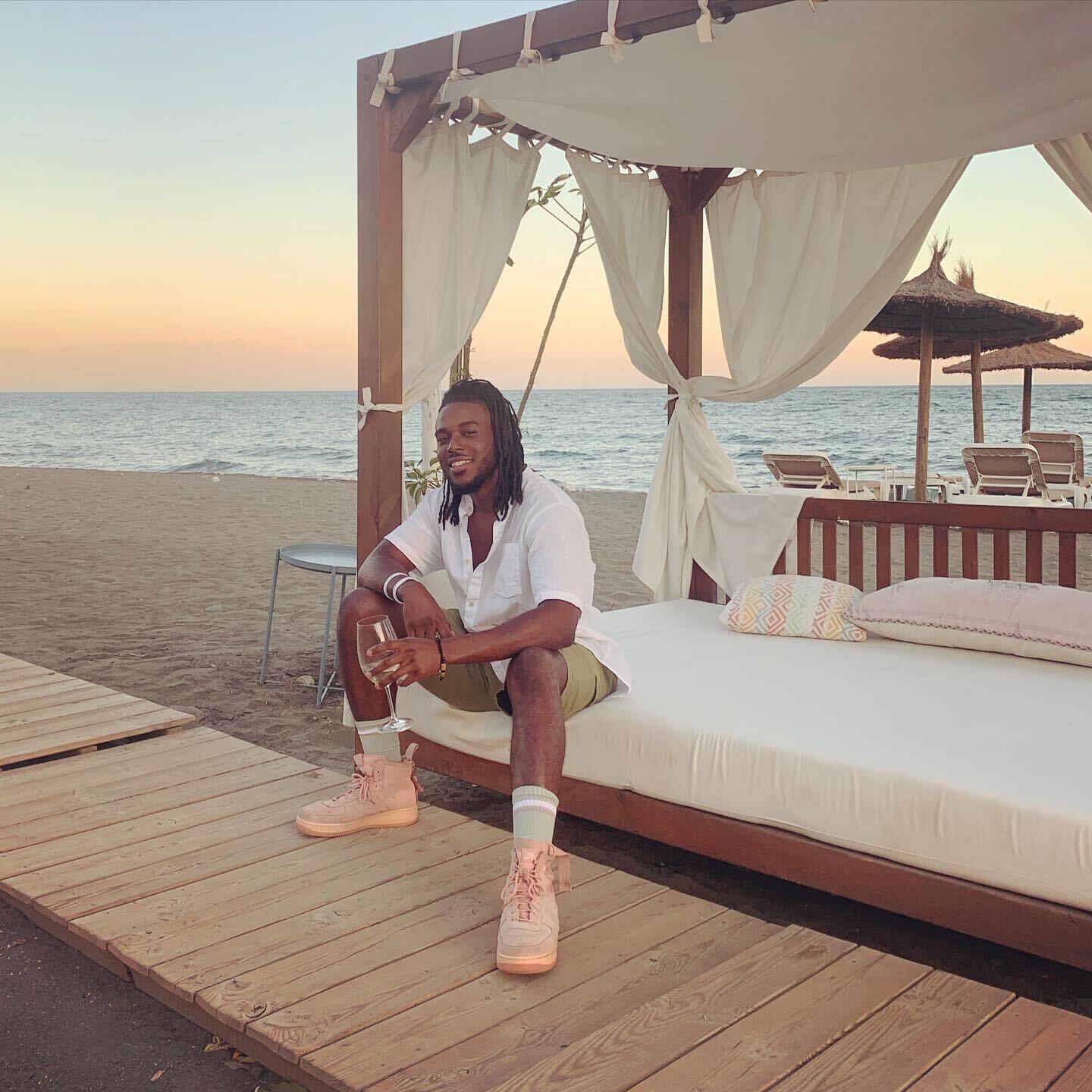 Black man sitting on a luxurious wooden beach bed with the calm spanish sea and gradient evening sky as a backdrop